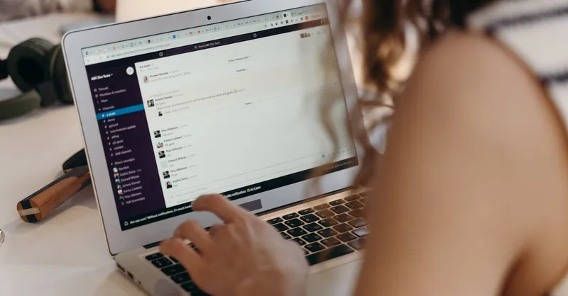 A businesswoman typing on a laptop in an office setting, using Slack for communication.