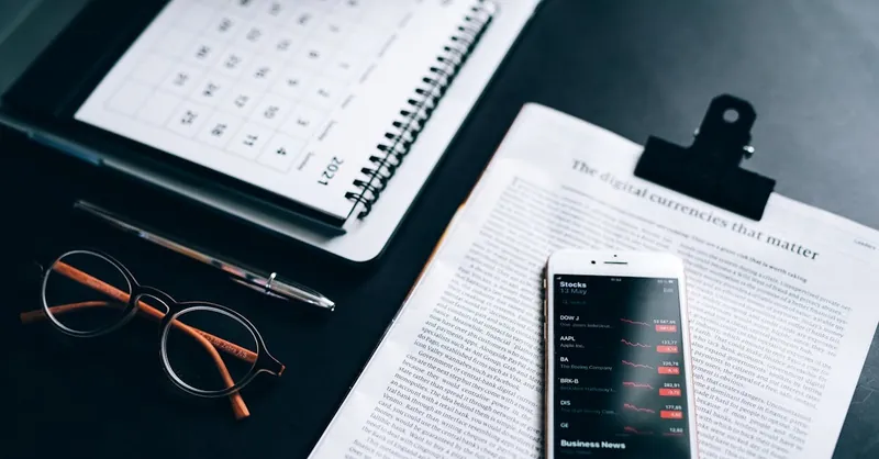 High angle shot of a desk setup with smartphone displaying stock market analytics, clipboard, pen, and glasses.