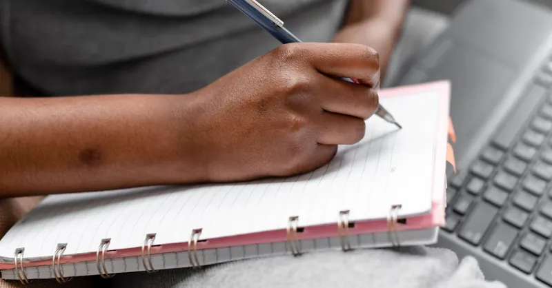 A person writing in a spiral notebook beside a laptop, focusing on note-taking and study.