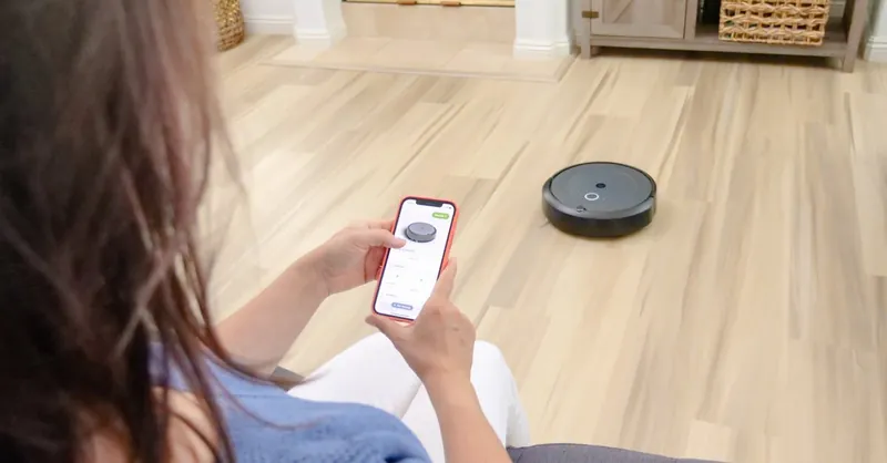 Overhead view of a woman controlling a robot vacuum using a smartphone app on a wooden floor.