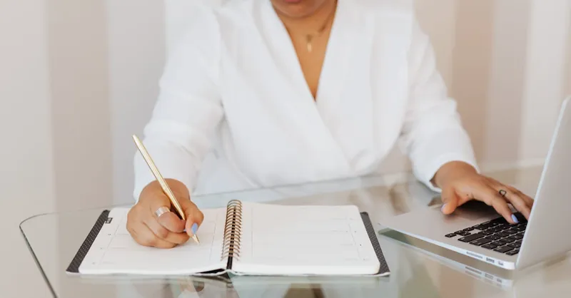 A focused professional woman planning her day with a notebook and laptop at a modern glass desk.