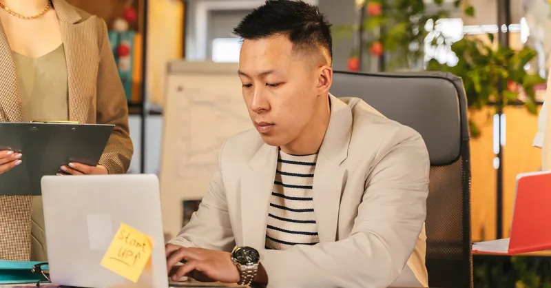 Asian man focusing on work in a modern office, engaged with a laptop, symbolizing a busy start-up environment.