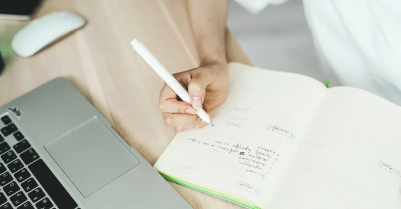 A hand writing in a notebook near a laptop, representing planning and freelancing.