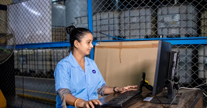 A woman in blue uniform operates a computer inside a factory warehouse, focusing on her task.