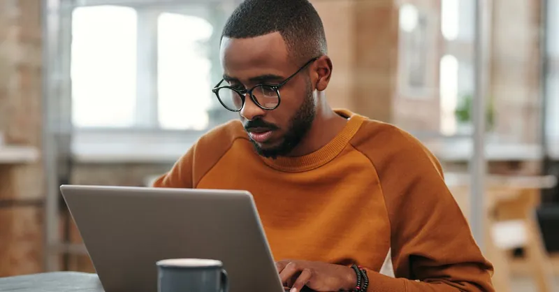 Young man with glasses works on laptop at a wooden table indoors. Warm, productive atmosphere.