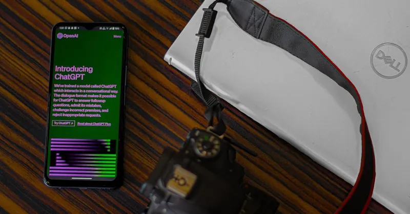 Smartphone with ChatGPT screen next to camera and laptop on wooden desk.