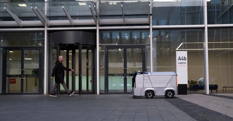 A modern office setting featuring a man and an autonomous delivery robot outside a glass-covered building.