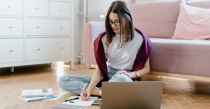A young woman studying indoors using a laptop and writing notes.