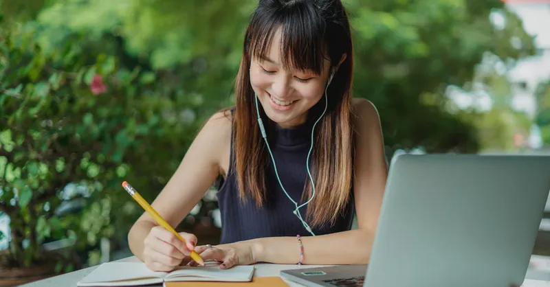 Positive young Asian female student with earphones writing in copybook while doing homework at table with laptop in street cafeteria