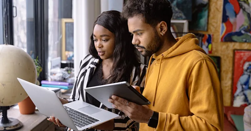 Two young adults collaborating using laptop and tablet indoors, showcasing teamwork and technology.