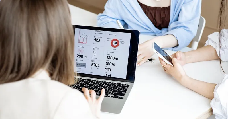 A group of women collaborating at a desk using a laptop and smartphone for business tasks indoors.