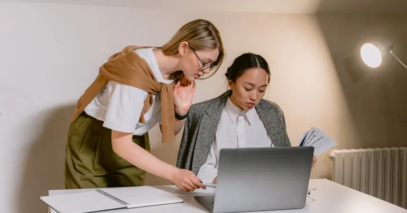 Two women collaborating at a desk with a laptop and documents in a modern office.