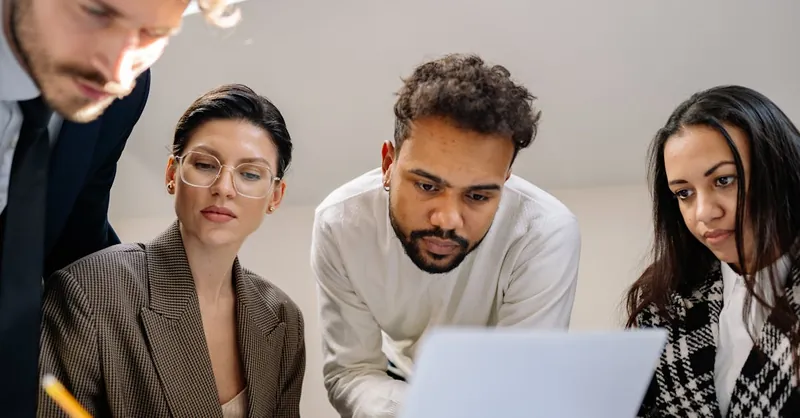 A diverse group of professionals collaborating on a project around a laptop in a modern office setting.